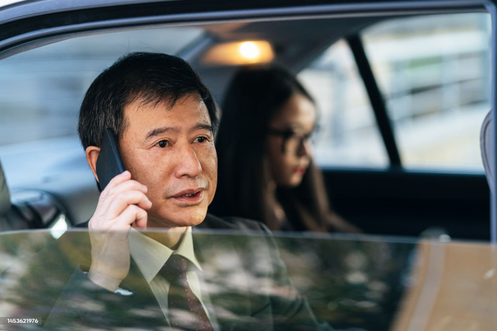 36214 Senior asian businessman talking on the phone while travelling together with young asian businesswoman on the backseat of a car on the way to a business meeting, watching through the window